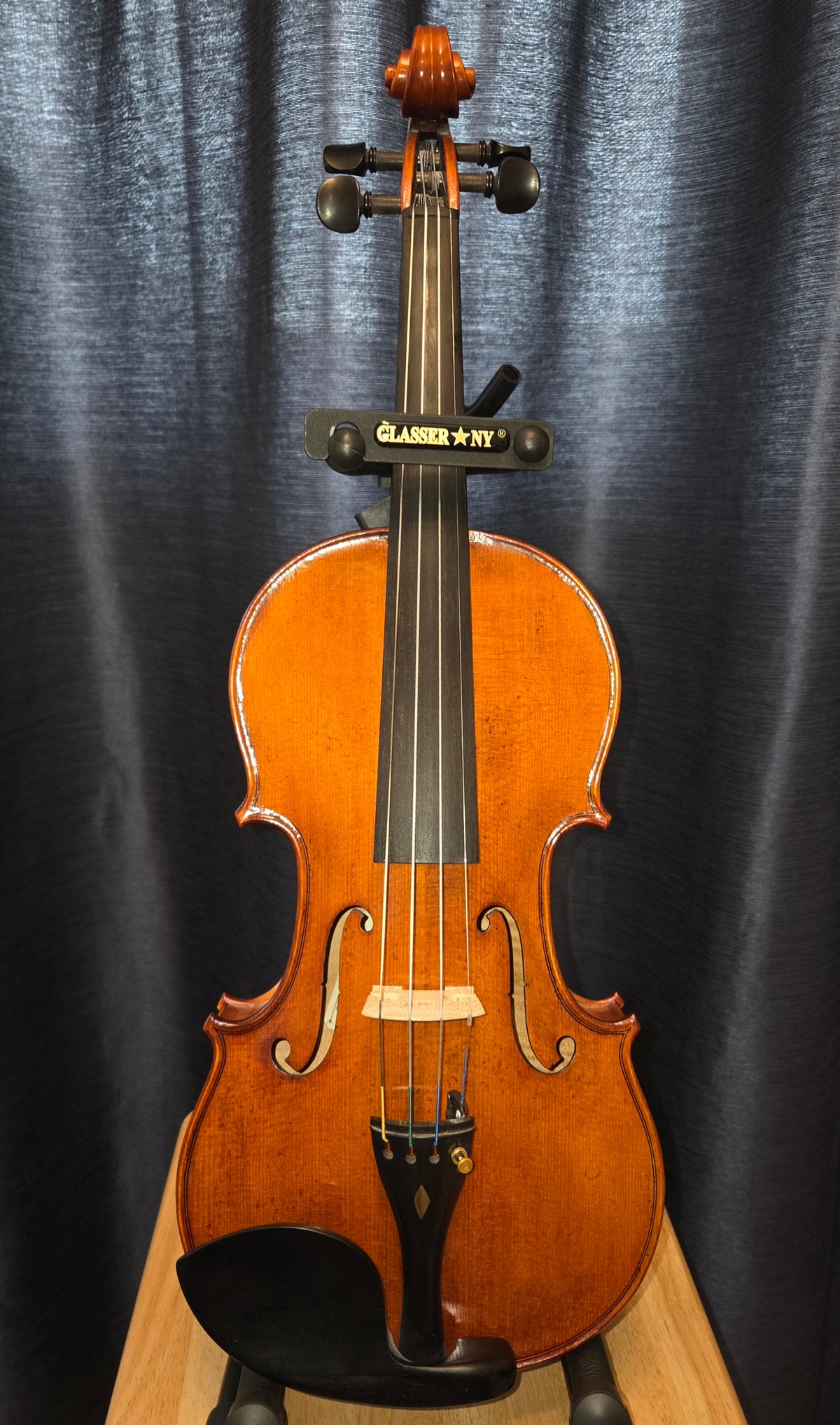 A classic wooden violin with a glossy orange-brown finish, displayed upright on a stand against a dark blue-gray curtain backdrop.